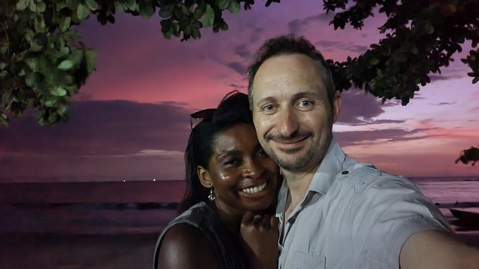 Un couple joue contre joue au premier plan, souriant, devant un ciel de coucher de soleil rose au-dessus de l'océan