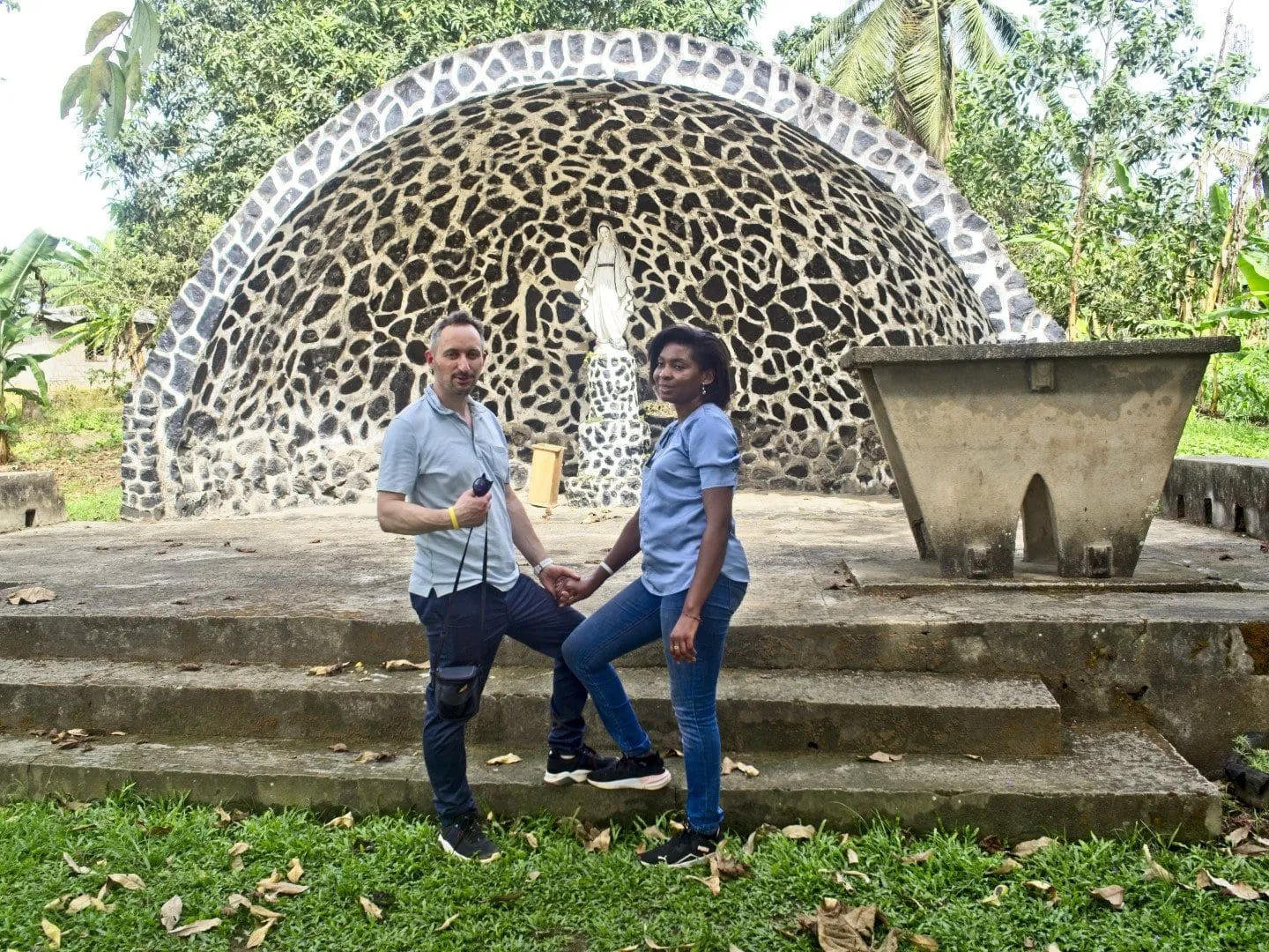 Une arche de pierres claires et sombres abritant une statue blanche, deux personnes devant