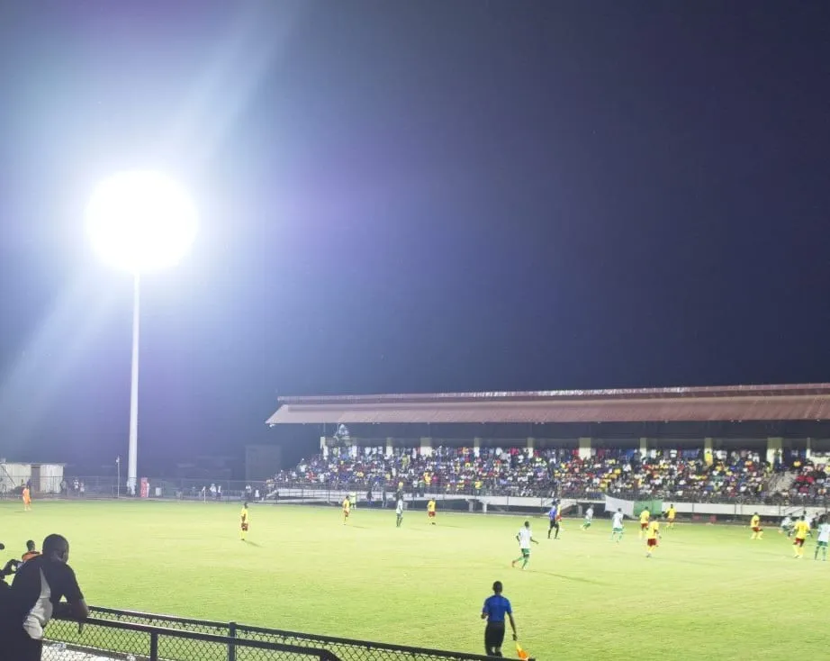 Un terrain de football éclairé par un projecteur, joueurs en action, tribune pleine au fond