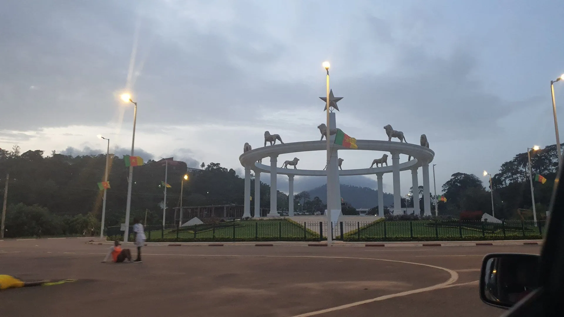 Une arche monumentale surmontée de statues de lions, éclairée, à un carrefour désert au crépuscule