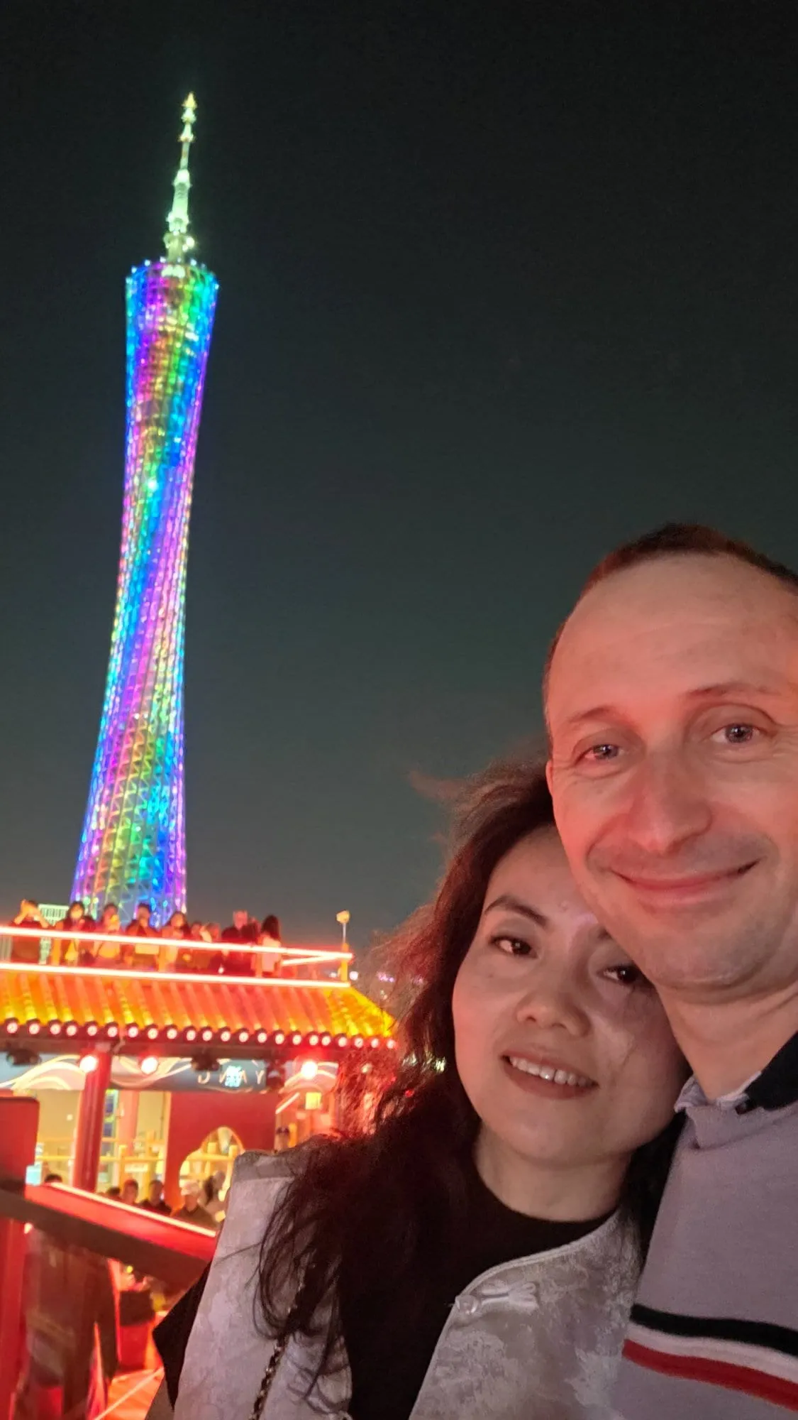 Selfie de 2 personnes souriantes devant la Canton Tower illuminée de nuit, la tour dressée derrière elles