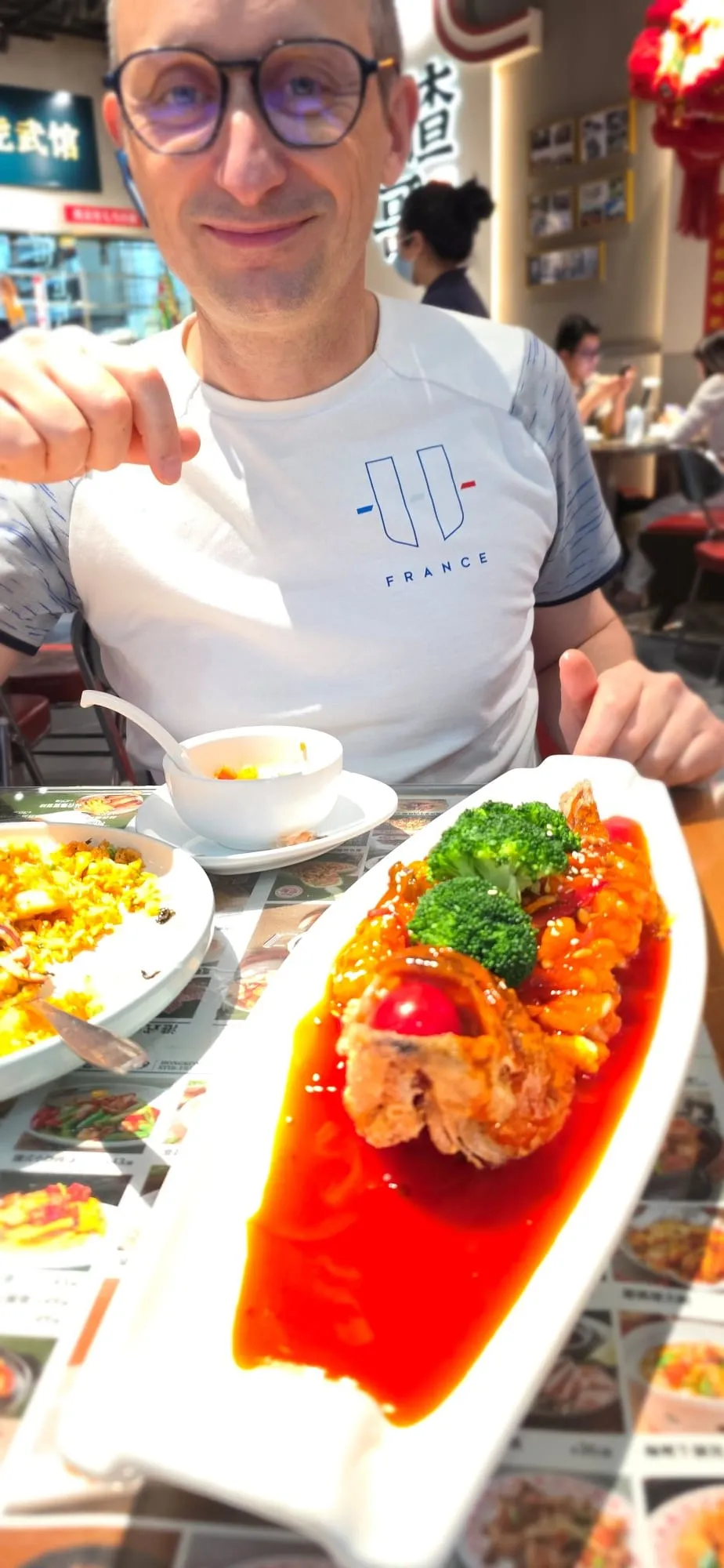 Un homme attablé présentant un long plat blanc où un poisson entier est nappé de sauce rouge