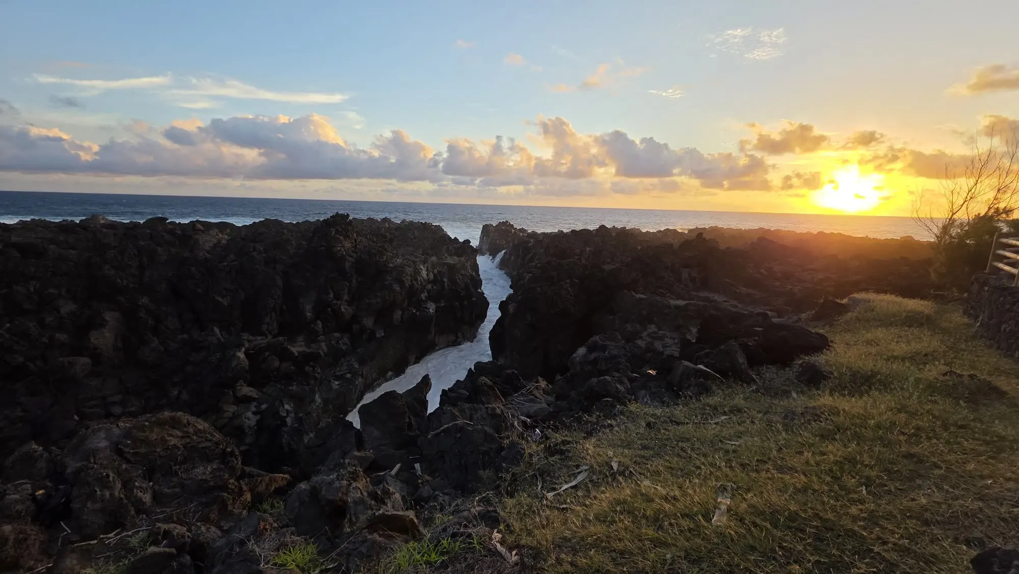 Une côte de lave noire descendant vers l'océan au coucher du soleil, traversée par un chenal d'eau