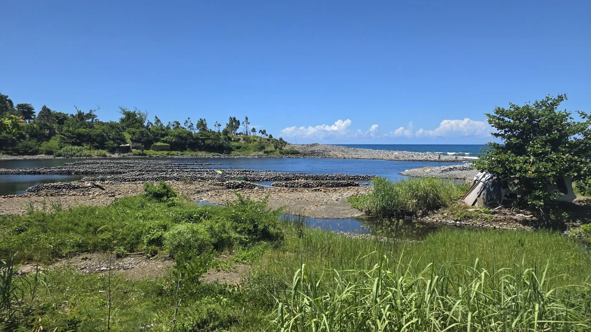 Une rivière à galets rejoignant l'océan entre deux rives végétales, ciel bleu
