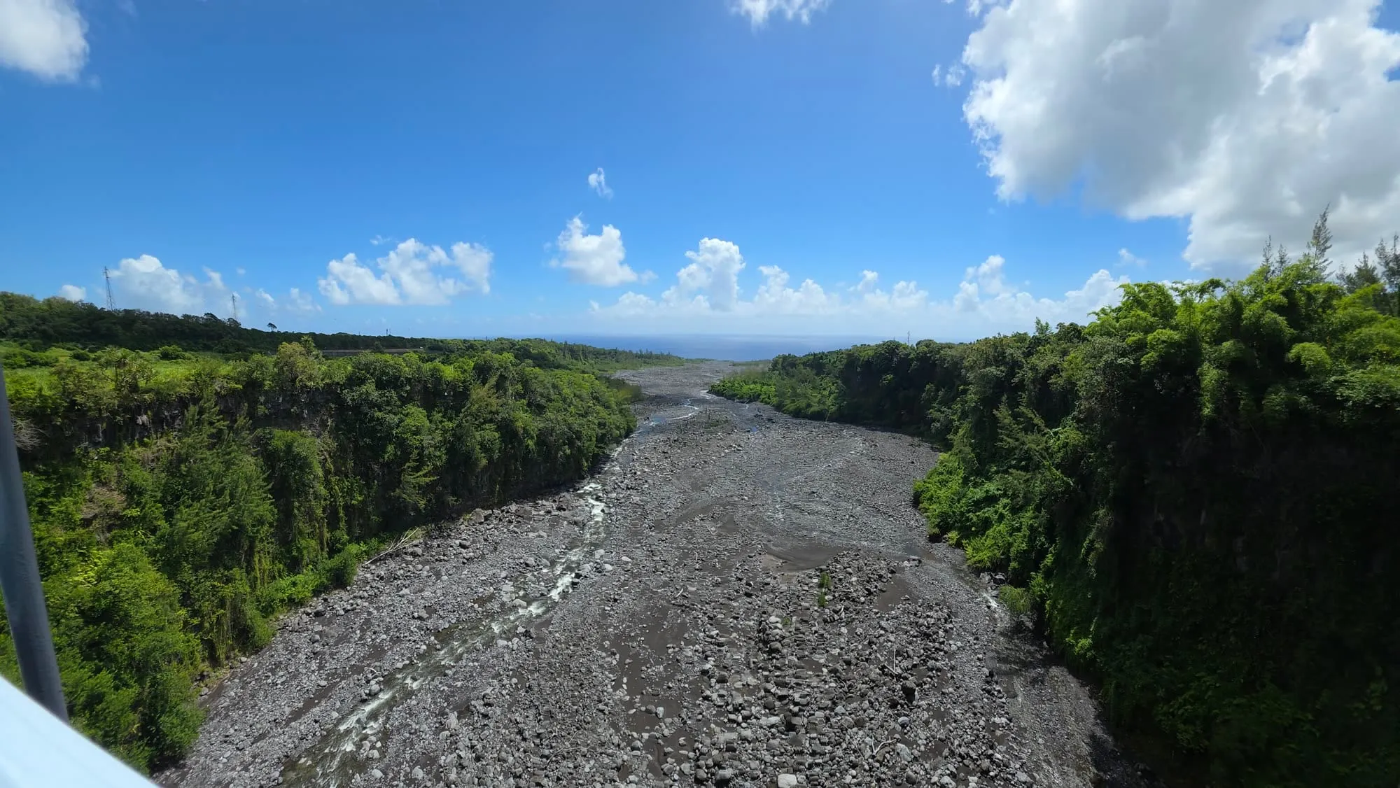 Un large lit de galets gris entre deux rives végétales, océan à l'horizon