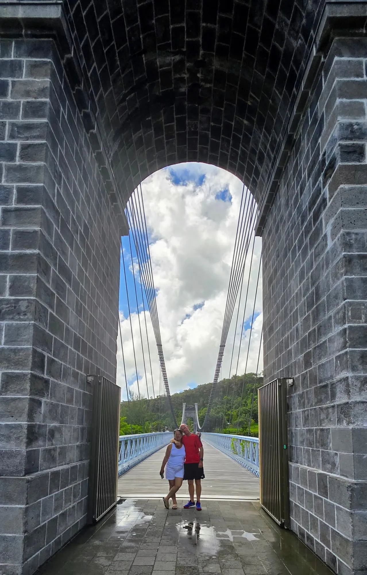 Une arche de pierre encadrant le tablier d'un pont suspendu, deux silhouettes engagées dessus