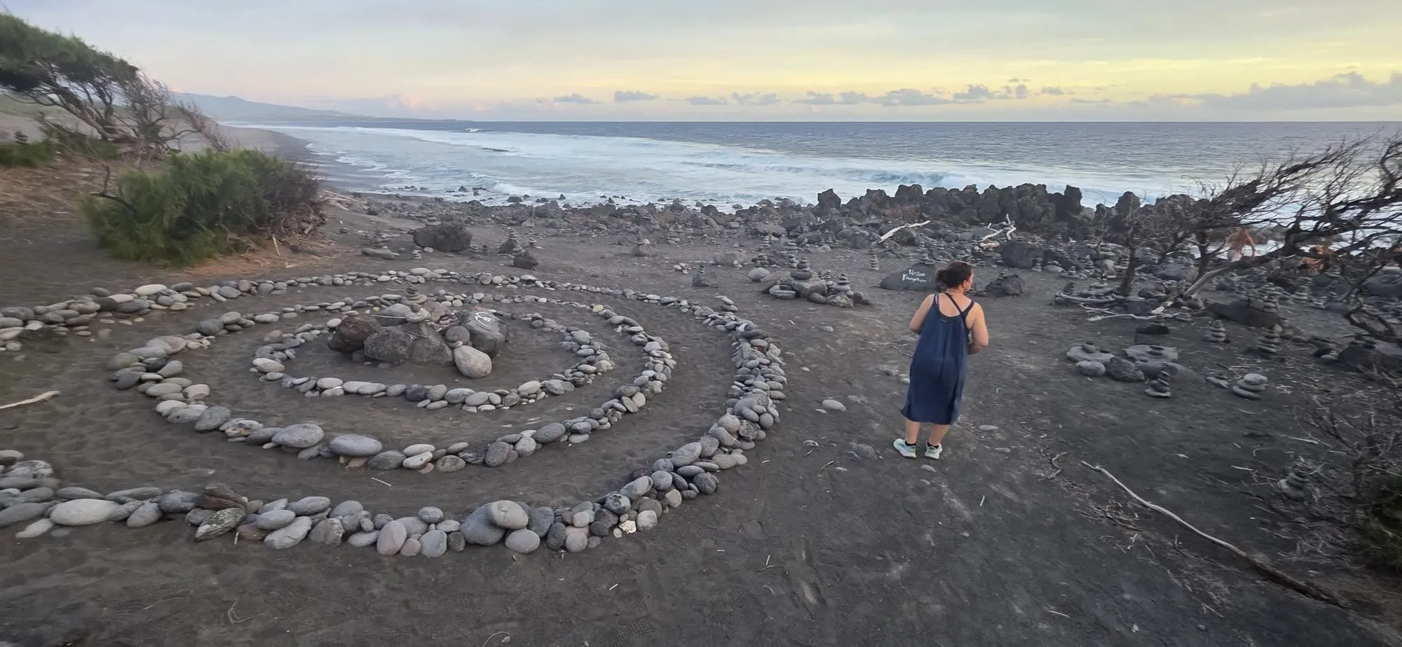 Des spirales de galets disposées sur une plage de sable noir, une silhouette au loin